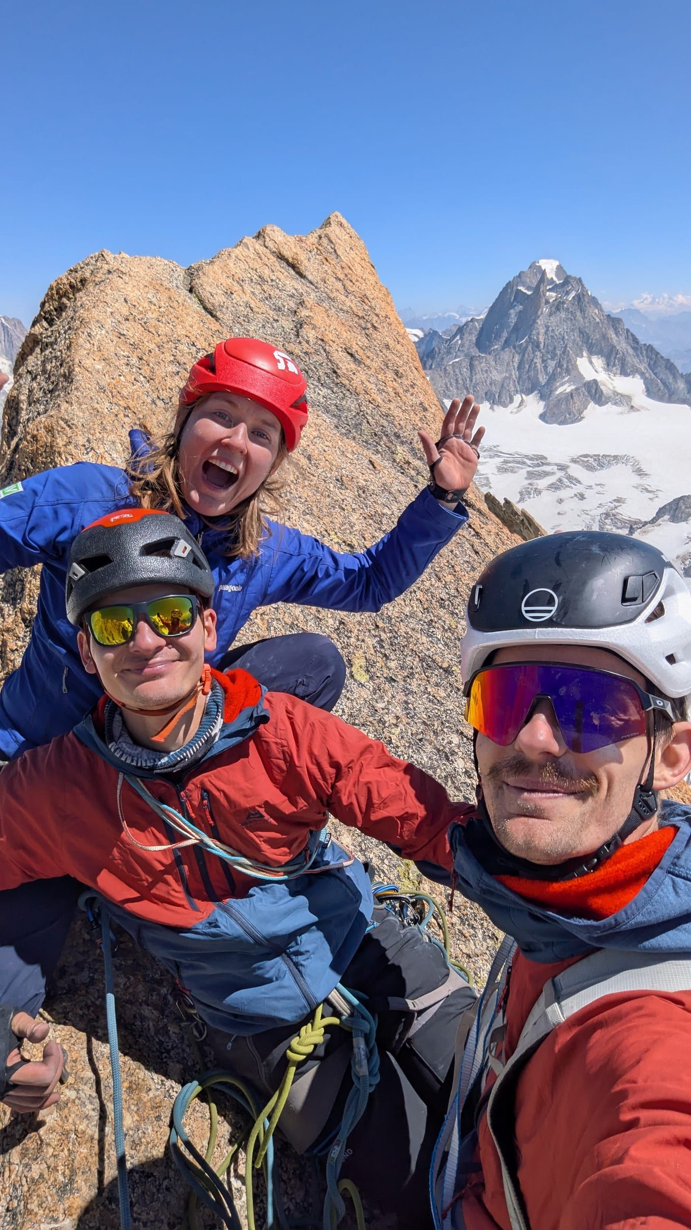 Drei Kletterer posieren lachend für ein Gipfelselfie, im Hintergrund Gipfel und Gletscher des Mont-Blanc-Massivs.